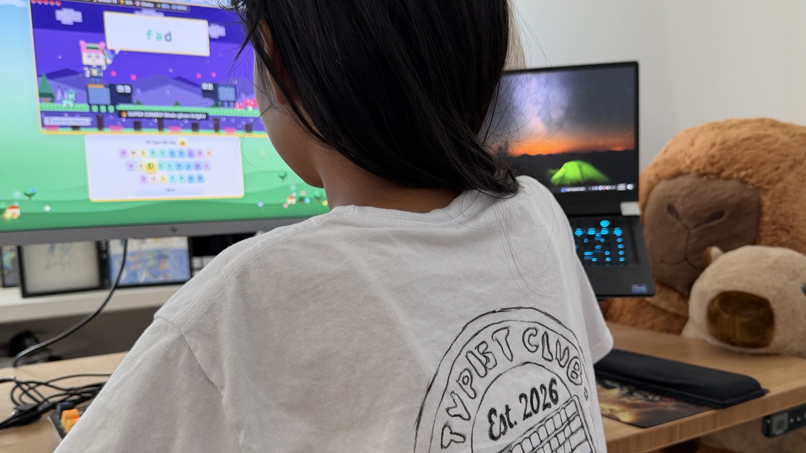 Working on Maya's Typing Adventure at a desk, game on screen, back of head and marker shirt visible.